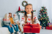 joyful girl holding christmas present and looking at camera while relatives sitting on background