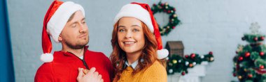 Noel Baba şapkalı bir kadının Noel 'de kocasını kucaklarken kameraya gülümseyişi.