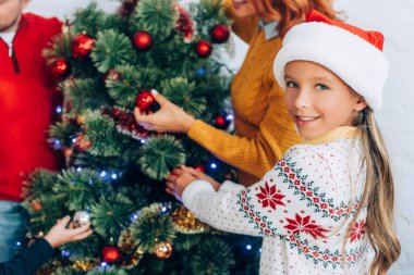 Noel Baba şapkalı mutlu kız aile ile birlikte Noel ağacı süslerken kameraya bakıyor.