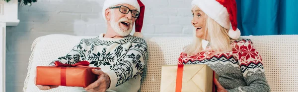 website header of excited senior couple in santa hats looking at each other while sitting on sofa with gift boxes