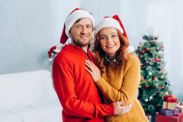 Felice Giovane Coppia Cappelli Babbo Natale Abbracciando Mentre Guardando Fotocamera — Foto Stock