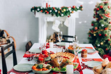 Masanın seçici odağı şenlikli akşam yemeği, Noel ağacı ve arka planda süslü ladin dalı olan şömine ile servis edilir.
