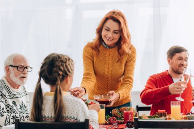 Redhead woman serving sauce on plate near family at festive table clipart