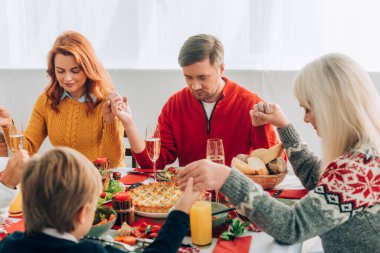 Selective focus of woman and man holding hands with family, sitting at table clipart