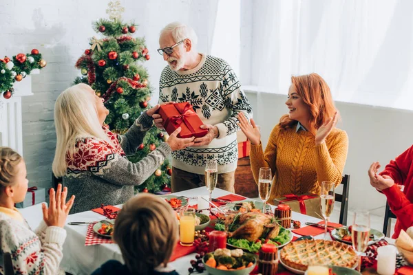 Glücklicher Großvater Grüßt Ehefrau Mit Geschenkbox Der Nähe Familie Applaudiert — Stockfoto