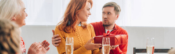 Панорамный снимок мужа, обнимающего счастливую жену с подарочной коробкой, сидящего рядом с семьей