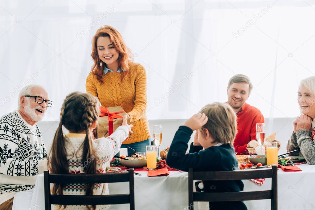 Mother greeting daughter with gift box, standing near festive table and family