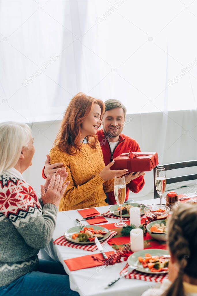 Husband embracing wife looking at gift box, sitting with family at festive table