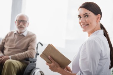 smiling social worker looking at camera while reading book to aged handicapped man on blurred background clipart