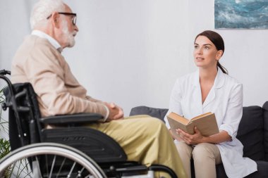 social worker in white coat reading book to aged disabled man in wheelchair on blurred foreground clipart