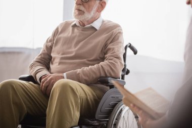Ön planda bulanık tekerlekli sandalyedeki engelliye kitap okuyan sosyal hizmet görevlisinin kırpılmış görüntüsü
