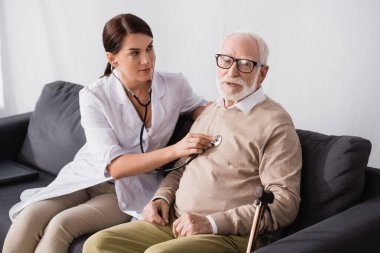 nurse examining aged man with stethoscope at home clipart