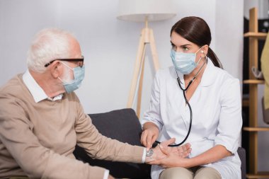 social worker in medical mask examining elderly man with stethoscope at home clipart
