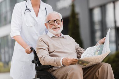 Sosyal hizmetler görevlisi tekerlekli sandalyedeki engelli adamla yürüyor elinde gazete tutuyor ve dışarıda kameraya gülümsüyor.
