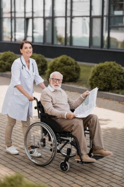 cheerful social worker looking at camera while walking with handicapped aged man with newspaper outdoors clipart