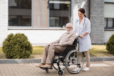 Gülümseyen sosyal hizmet görevlisi dışarıda tekerlekli sandalyedeki engelli bir adamla yürüyor.