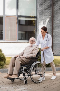 Neşeli sosyal hizmet görevlisi dışarıda tekerlekli sandalyede yaşlı engelli bir adamla yürüyor.