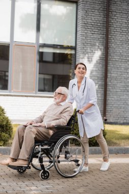 Tekerlekli sandalyedeki engelli adamla yürürken kameraya gülümseyen sosyal hizmet görevlisi.
