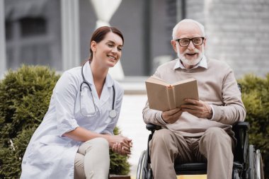 Yaşlı engelli bir adamın yanındaki neşeli yaşlı hemşire dışarıda tekerlekli sandalyede kitap okuyor.