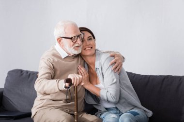 Minnettar yaşlı adam, evde koltukta otururken yaşlı hemşireye sarılıyor.