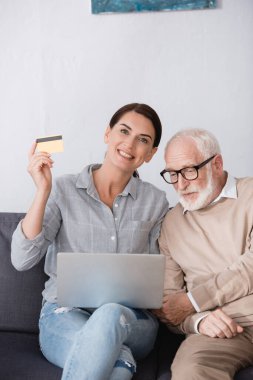 cheerful woman holding laptop and credit card while sitting near elderly father clipart
