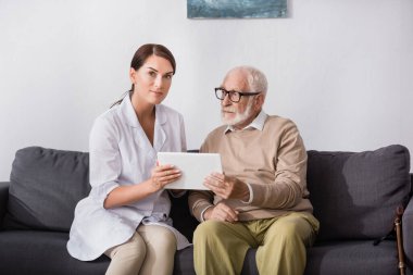 brunette social worker showing digital tablet to elderly man on sofa at home clipart