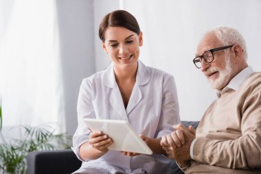 Gülümseyen sosyal hizmetler görevlisi yaşlı bir adama tablet gösteriyor ve elleri sımsıkı oturuyor.