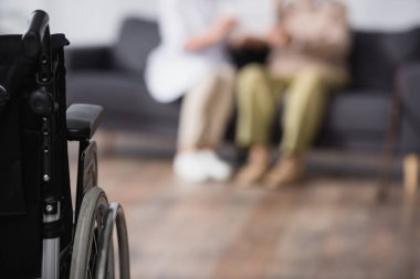 Tıbbi asistan ve yaşlı adam evdeki koltukta oturuyor tekerlekli sandalyenin yanında bulanık ön planda.