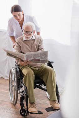Sosyal hizmet görevlisinin yanında tekerlekli sandalyede gazete okuyan engelli bir adam.