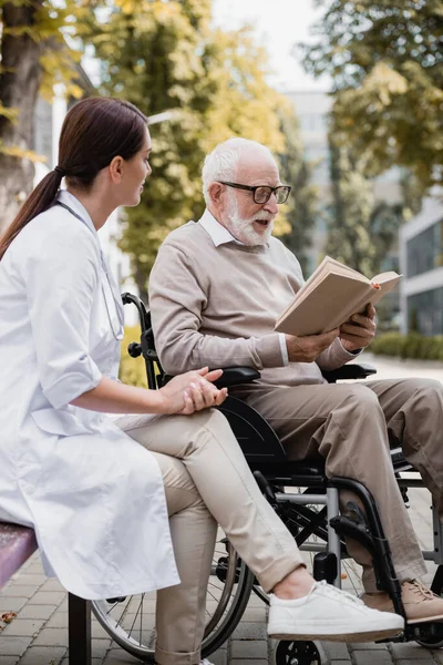 Geriatryczna Pielęgniarka Siedzi Pobliżu Wieku Niepełnosprawny Człowiek Czytanie Książki Zewnątrz — Zdjęcie stockowe