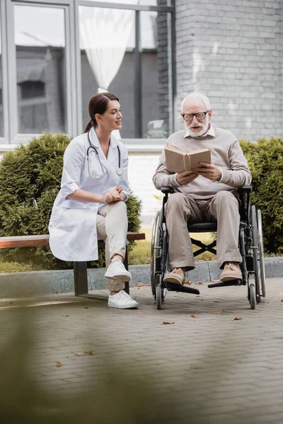 Handicapped Aged Man Reading Book Wheelchair Social Worker Sitting Bench — Stock Photo, Image