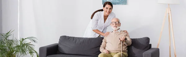 geriatric nurse and aged man smiling at camera together at home, banner