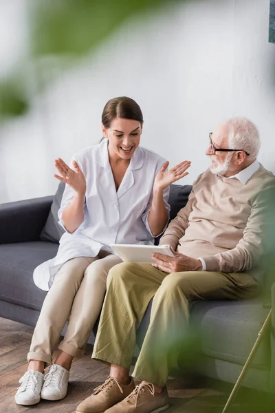 excited geriatric nurse sitting with open arms near elderly man and digital tablet on blurred foreground