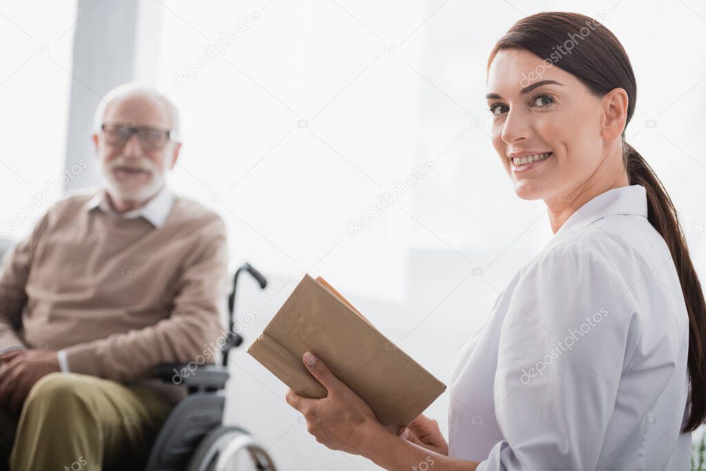 smiling social worker looking at camera while reading book to aged handicapped man on blurred background