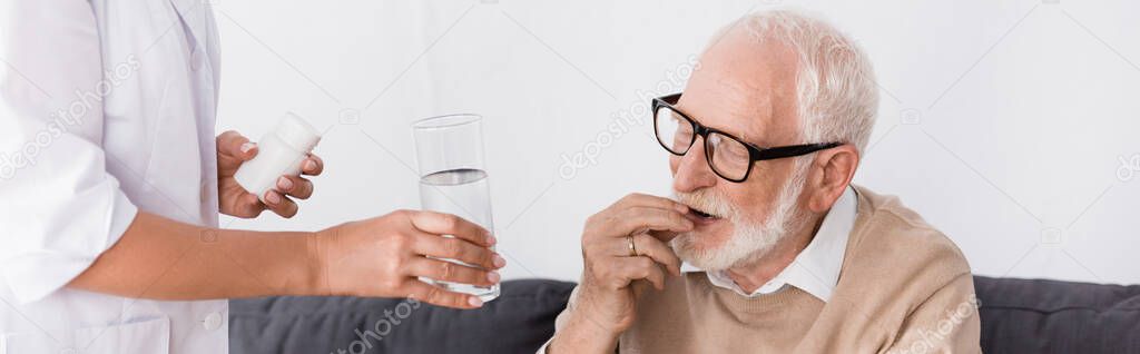 geriatric nurse holding pills and water near aged man, banner