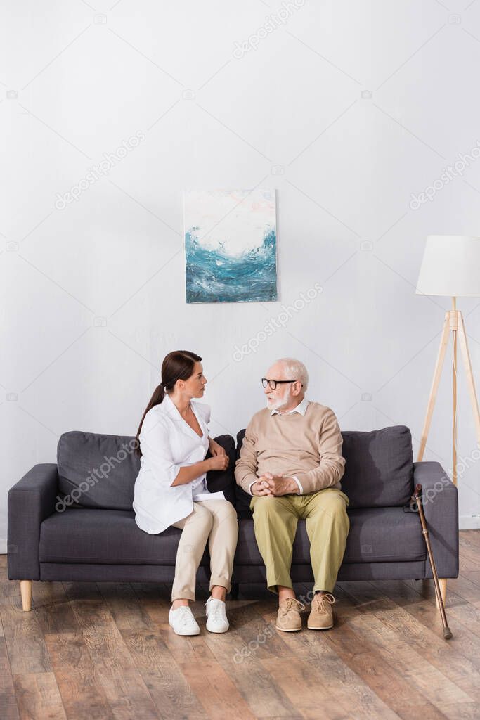 aged man in eyeglasses talking to geriatric nurse while sitting on sofa at home 