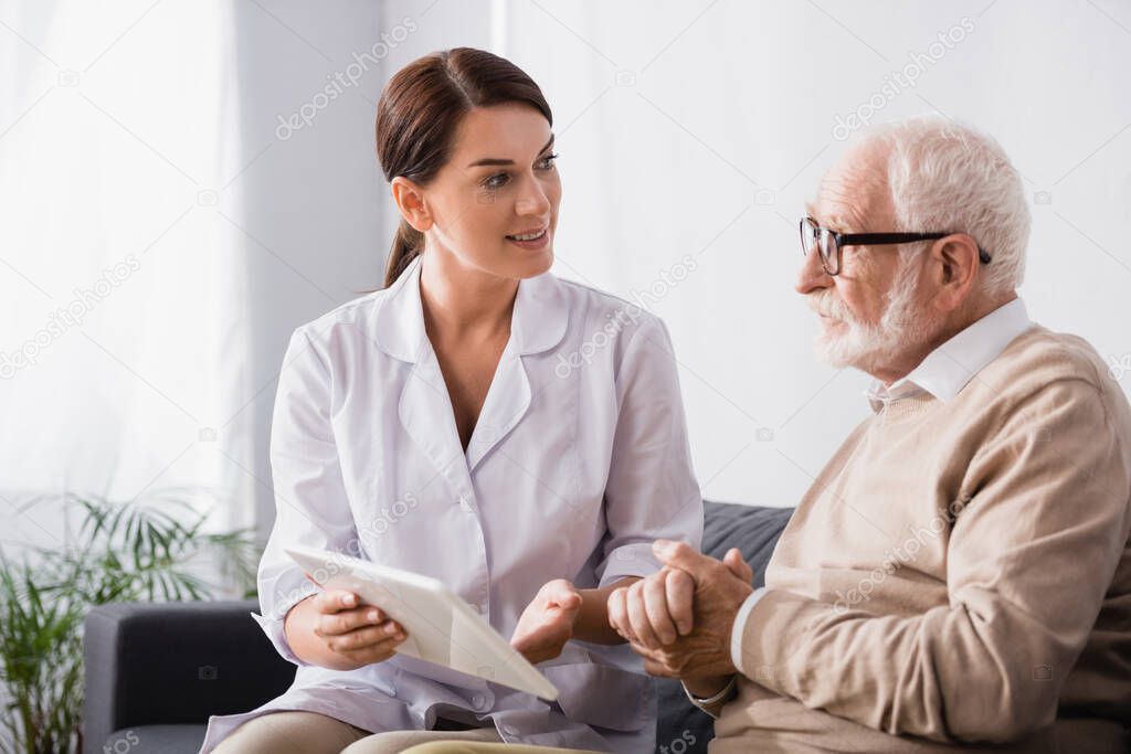 social worker holding digital tablet and talking to aged man sitting with clenched hands