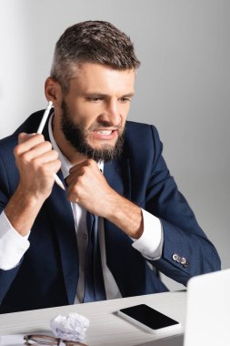 Akıllı telefonun yanında kırık kalem tutan gergin bir işadamı. Ön planda kümelenmiş kağıt ve gözlük var. 