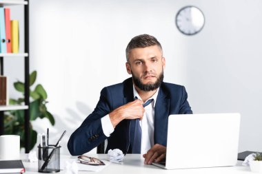 Businessman looking at camera while suffering from heat near stationery and laptop on blurred foreground on working table  clipart
