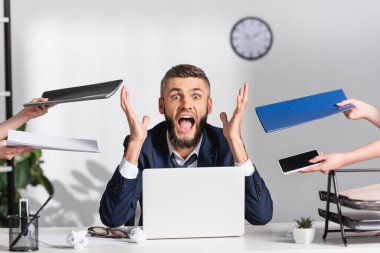 Stressed businessman screaming near colleagues with paper folders and gadgets in office  clipart