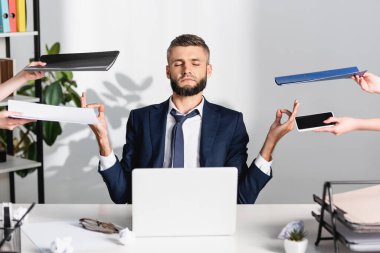 İş adamı, ofisteki bulanık ön planda doküman ve aletlerle meslektaşlarının yanında meditasyon yapıyor. 