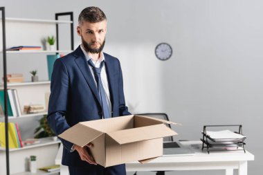 Kovulan işadamı, arka planda bulanık ofis masası olan karton bir kutu tutuyor. 