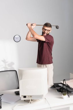 Bilgisayarın yanında golf sopası tutan stresli iş adamı ve ofisin ön planında bulanık kağıtlar var. 