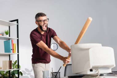 Çığlık atan iş adamı bilgisayar monitörünü beyzbol sopasıyla dövüyor. Ofis masasında bulanık bir ön planda. 