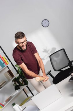 Stresli iş adamı ofis masasında beyzbol sopasıyla dizüstü bilgisayarı dövüyor 