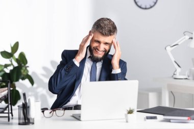 Dizüstü bilgisayarın yanında baş ağrısı çeken bir iş adamı ve çalışma masasındaki kağıtlar. 