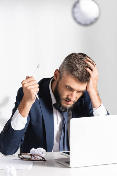 Напряженный бизнесмен с рукой рядом с головой держит сломанный карандаш рядом с ноутбуком, очки и сгущенную бумагу на размытом переднем плане 