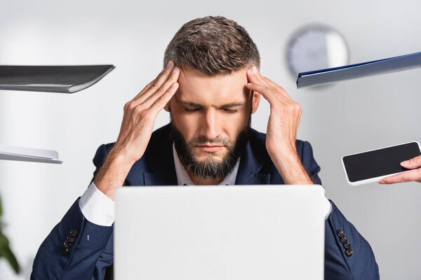 Tired businessman touching head near paper folders and gadgets on blurred foreground in office