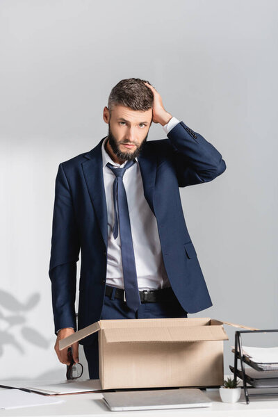 Dismissed businessman looking at camera near carton box, laptop and stationery on blurred foreground in office 
