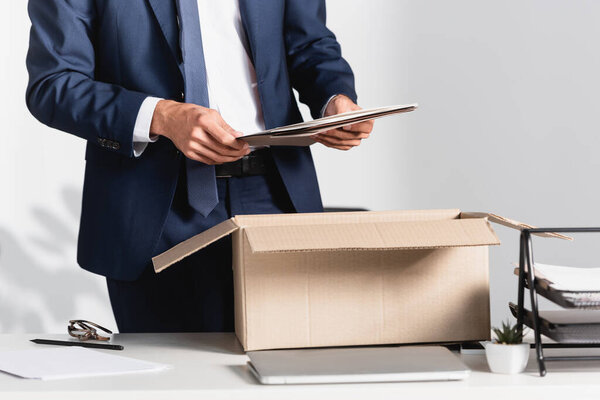 Cropped view of dismissed businessman holding paper folder near laptop and cardboard box on blurred foreground 